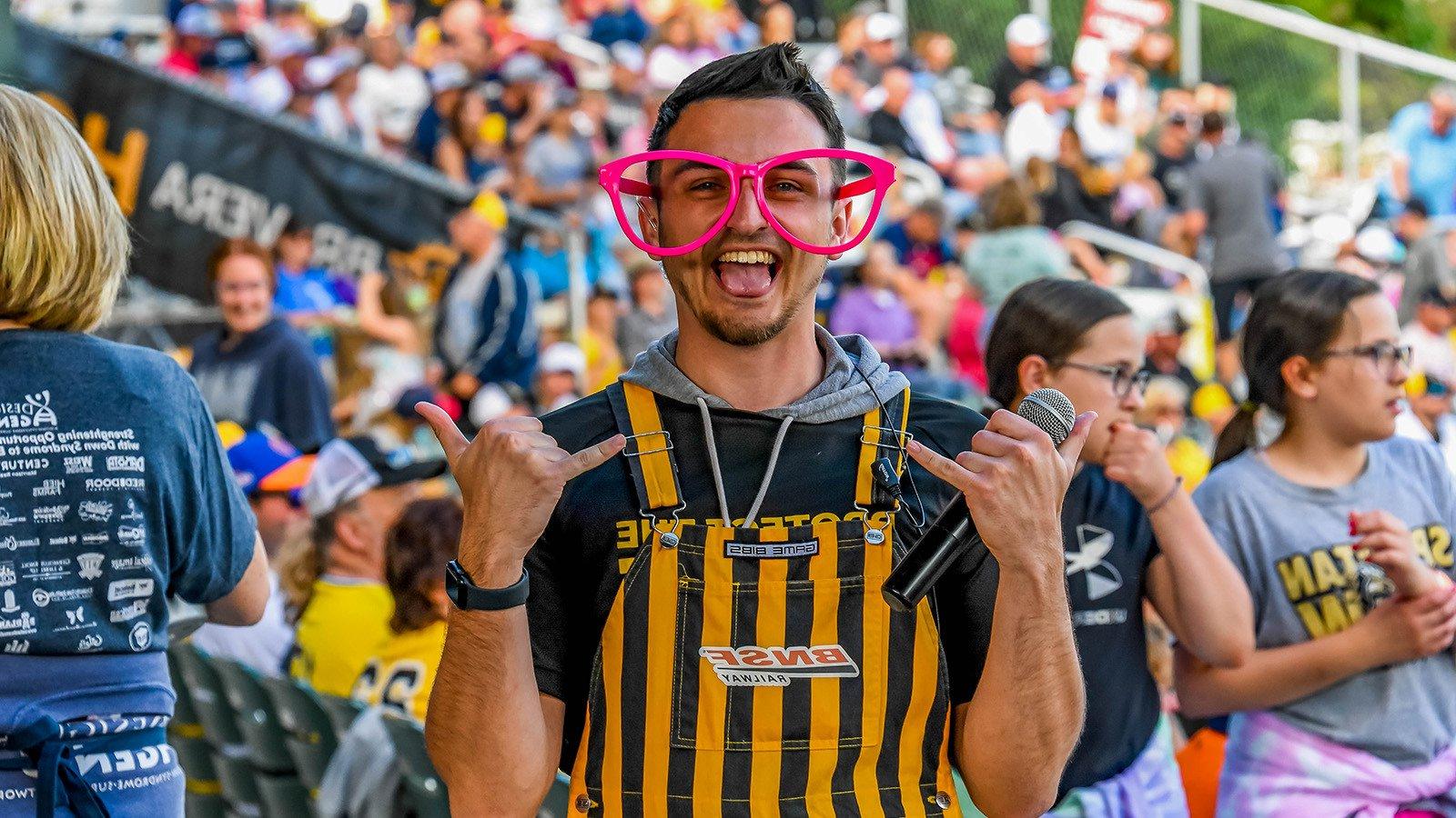 A man standing and looking excited while holding a microphone in oversized pink glasses and yellow and black striped overalls in the foreground of a large crowd at a stadium.